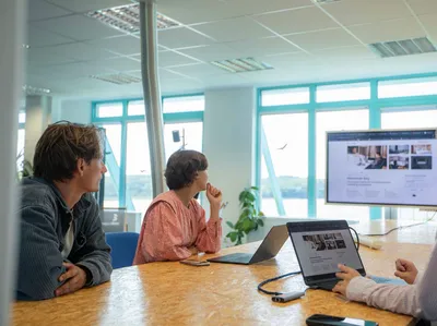 Team meeting at a wooden table with laptops, everyone looking at a screen in front of large windows