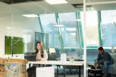 Glass-walled workspace in the coworking area, a woman working at a desk and a man with a laptop on the sofa