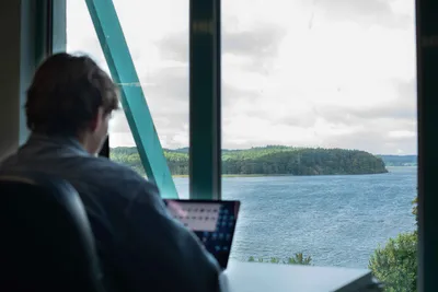 Man working on a laptop in front of a large window overlooking the lagoon and wooded shores